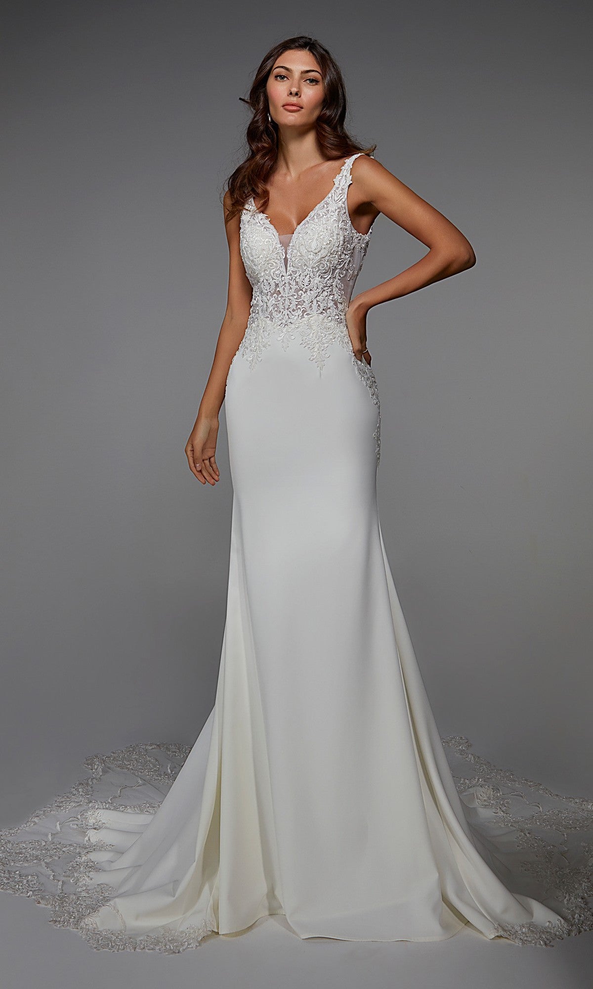 A woman wears the Alyce 7027 long ivory dress, featuring a sleeveless fitted bodice with intricate lace embroidery and a flowing train, standing against a plain gray background.